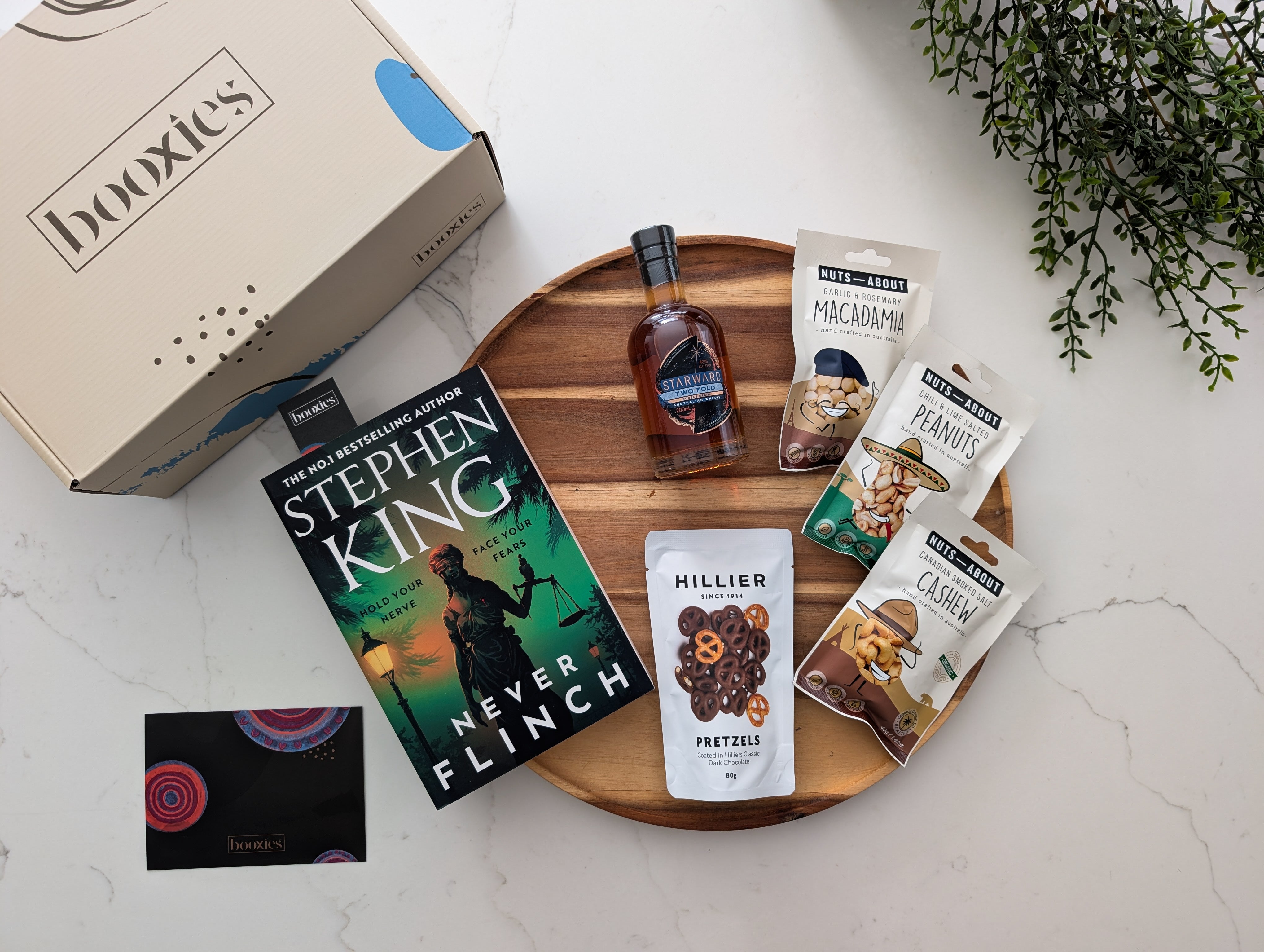 Gourmet gift set with 'Never Flinch' book, snacks, and a bottle of whisky on a white surface.