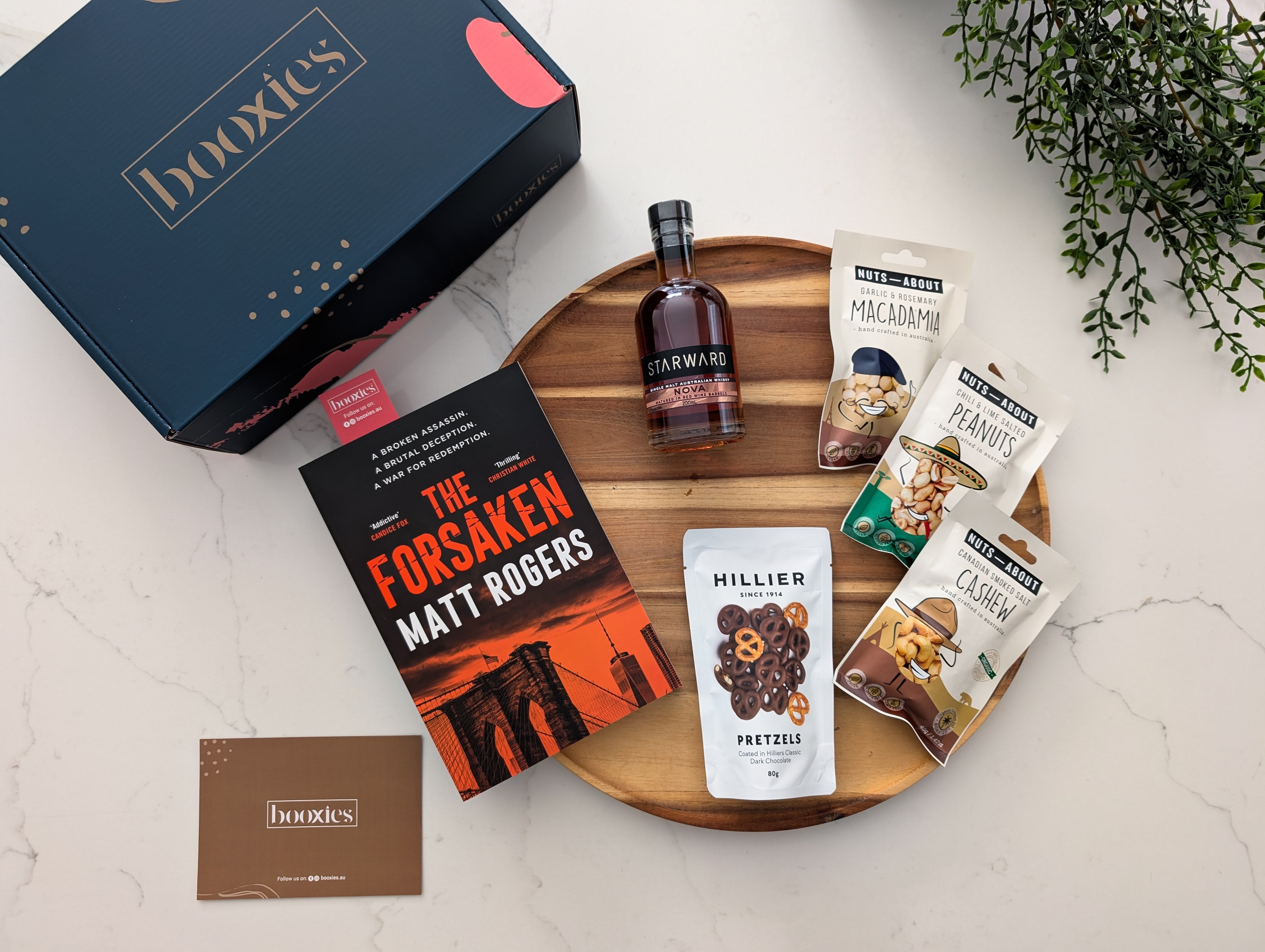 Book and snacks on a wooden tray with 'booxxies' branding featuring the forsaken by Matt Rogers