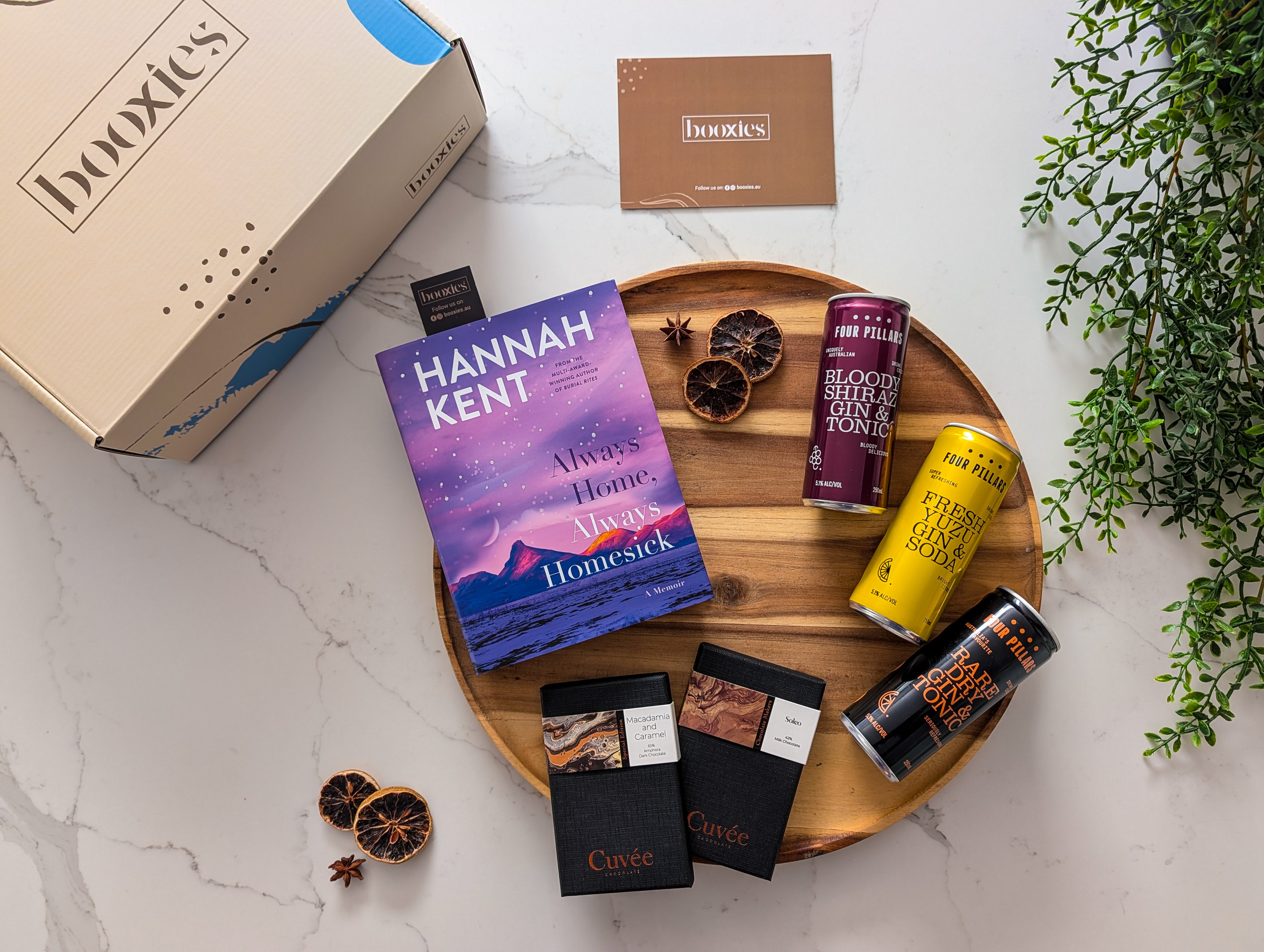 Hannah Kent book on a wooden tray with chocolate bars and a box labeled 'booxties' and gin rtd from four pillars on a marble surface.
