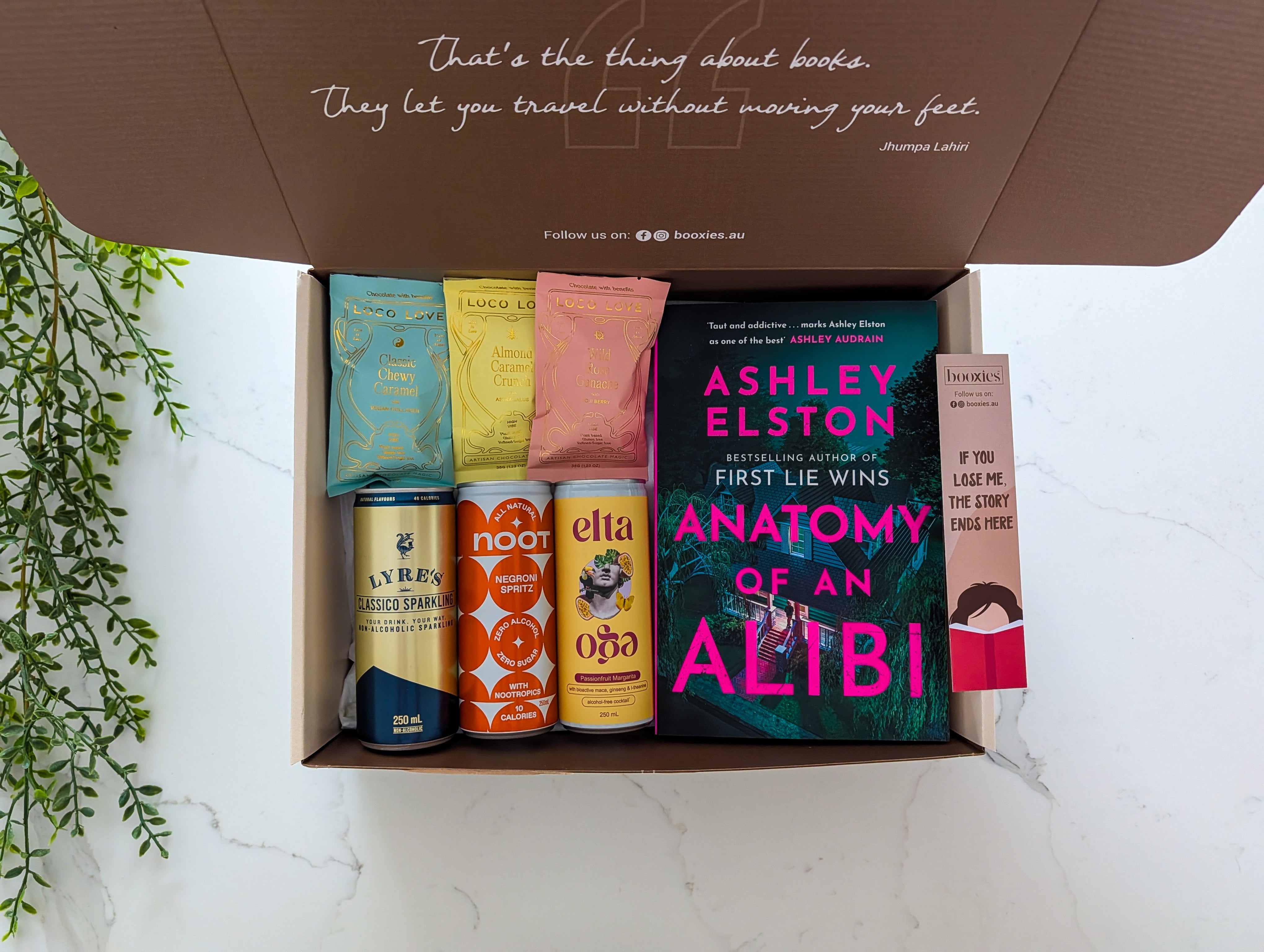 Open box containing books and drinks on a white surface with a plant on the side.