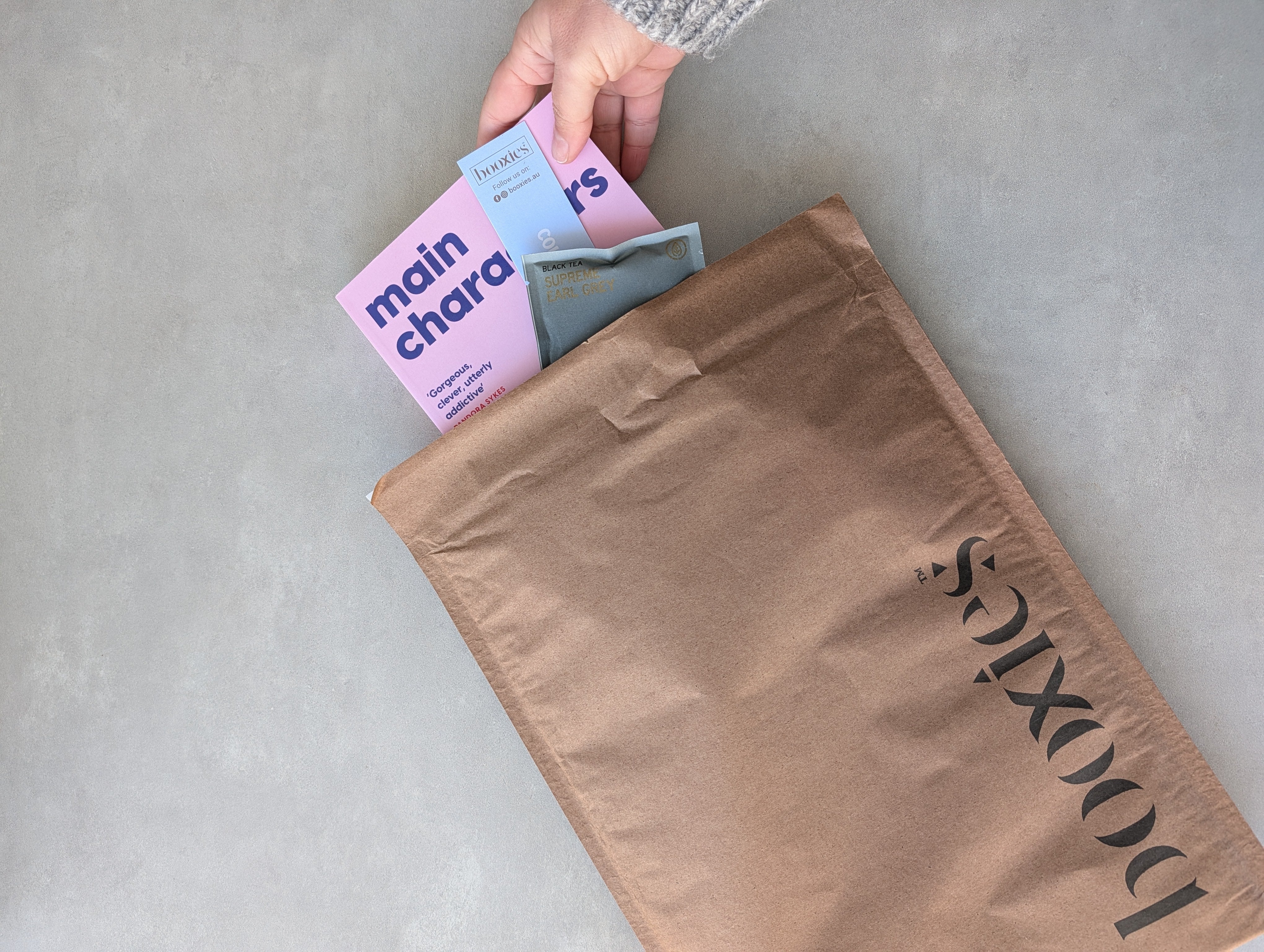A brown Booxies branded mailer envelope on a gray concrete floor, with a hand reaching in to pull out a pink book and other items.