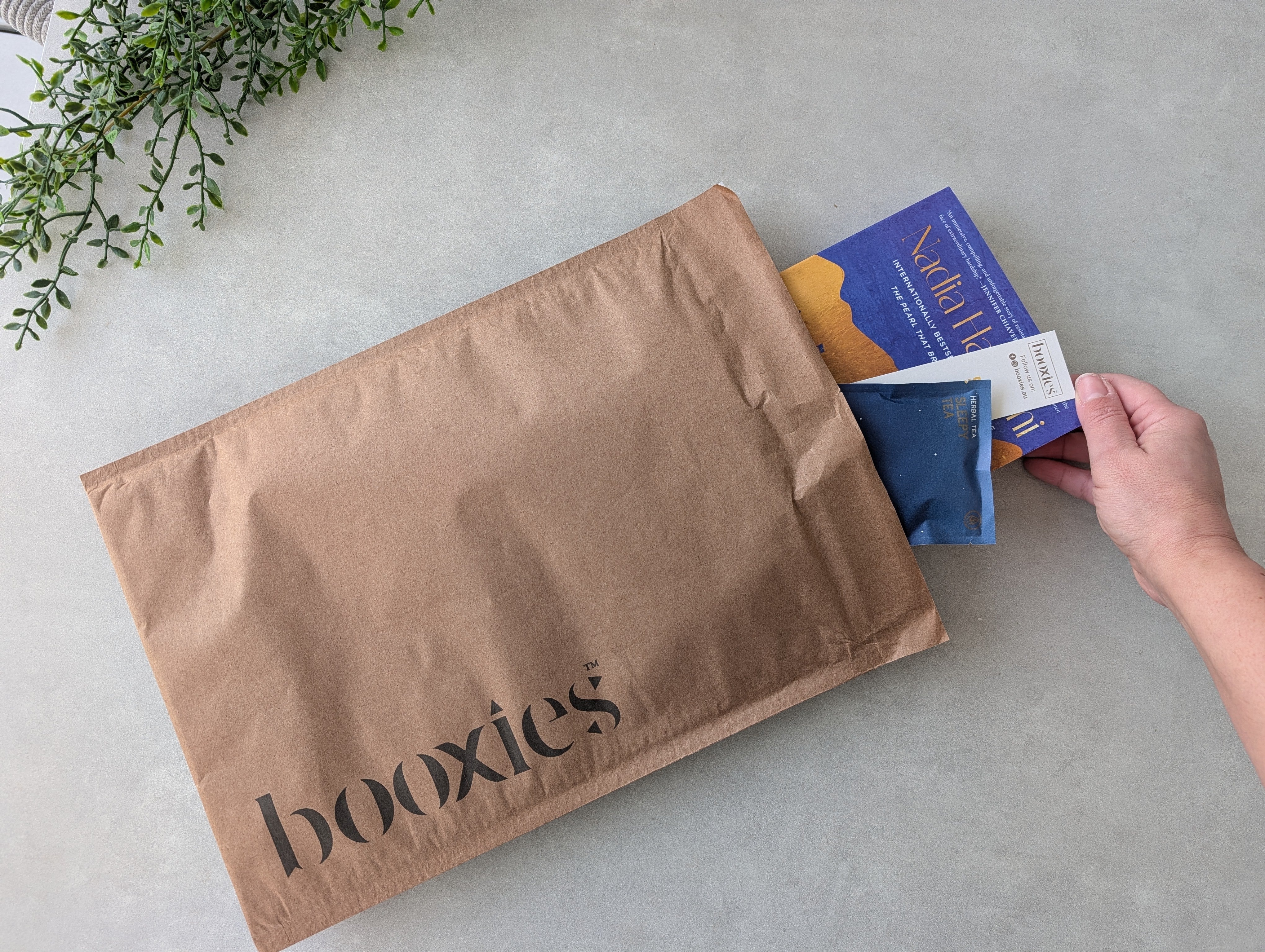 essential mystery booxies showing a hand with the book of the month and a bookmark and tea pyramids