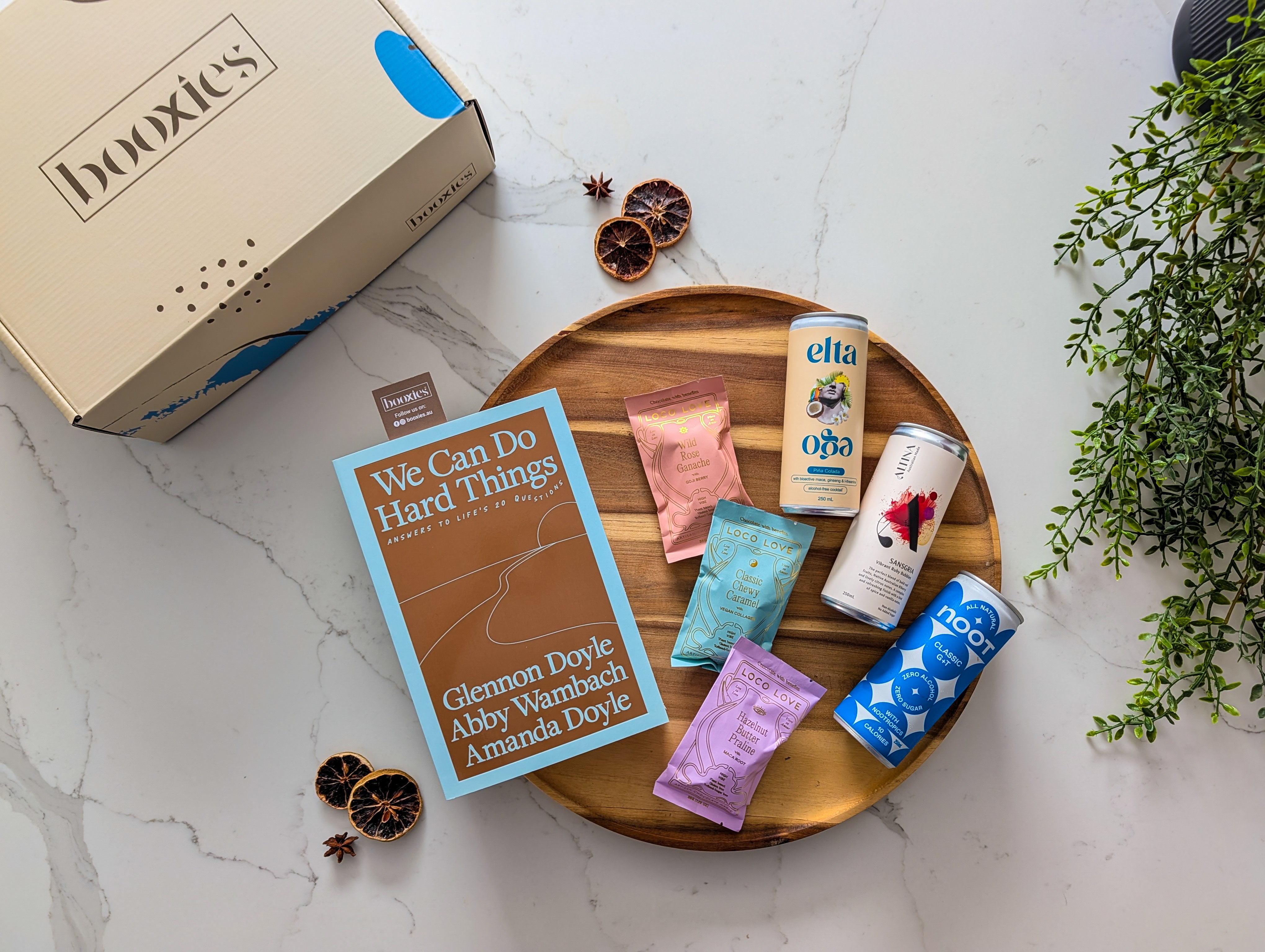 Assorted loco love chocolate and non-alcoholic drinks on a wooden tray with a 'booxies' branded box and a new book in the background. making up the Zero Edit curated booxies