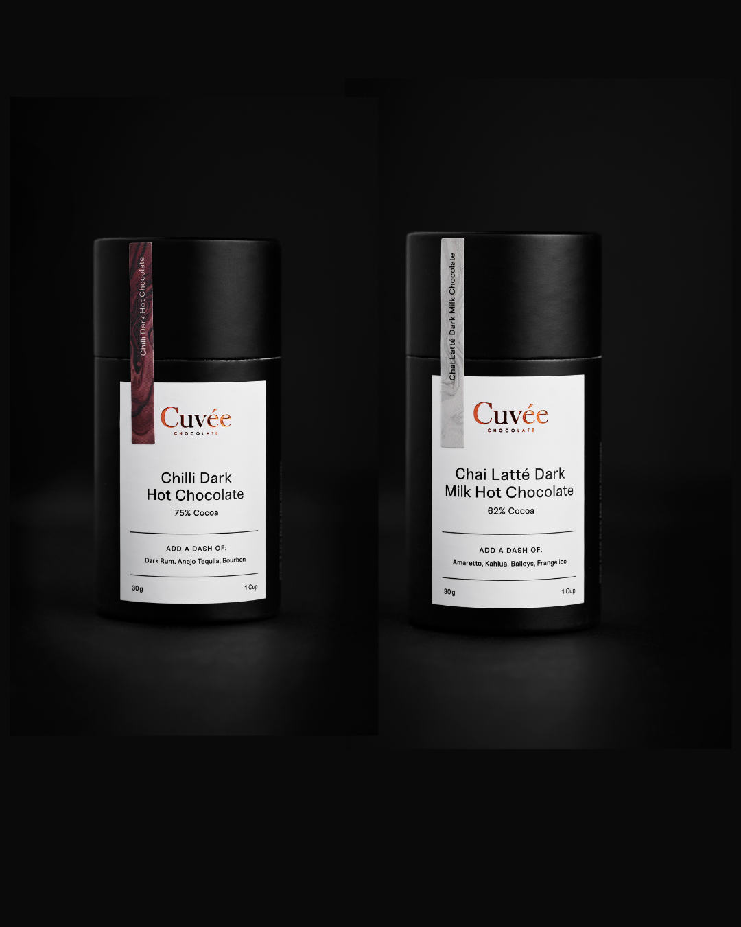 Two black cylindrical containers of Cève Chilli Dark Hot Chocolate and Chai Latè Dark Milk Hot Chocolate on a dark background.