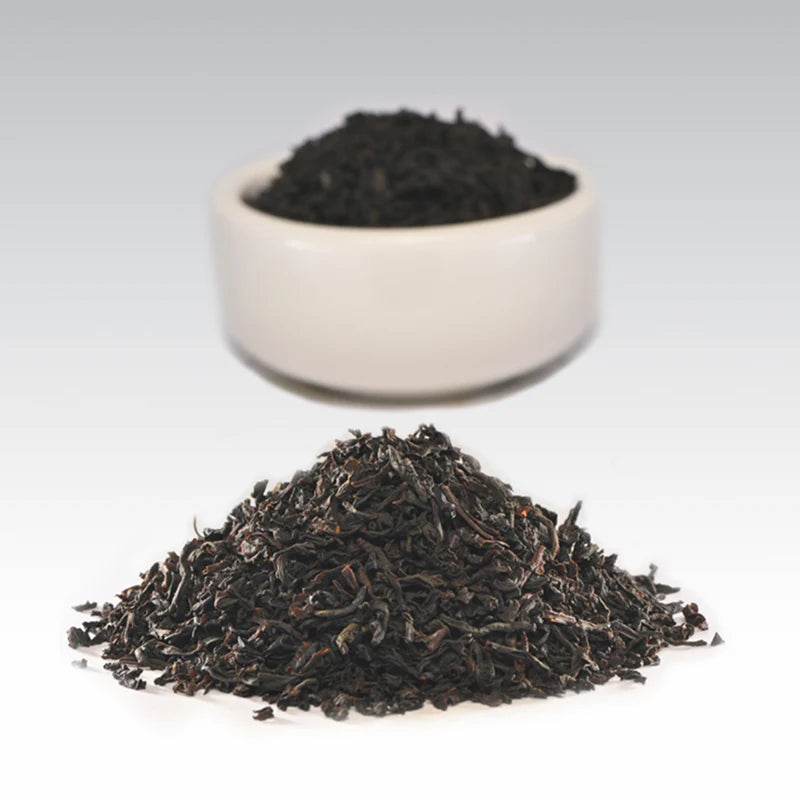 Dried black tea leaves with a small white bowl containing more tea leaves on a light gray background