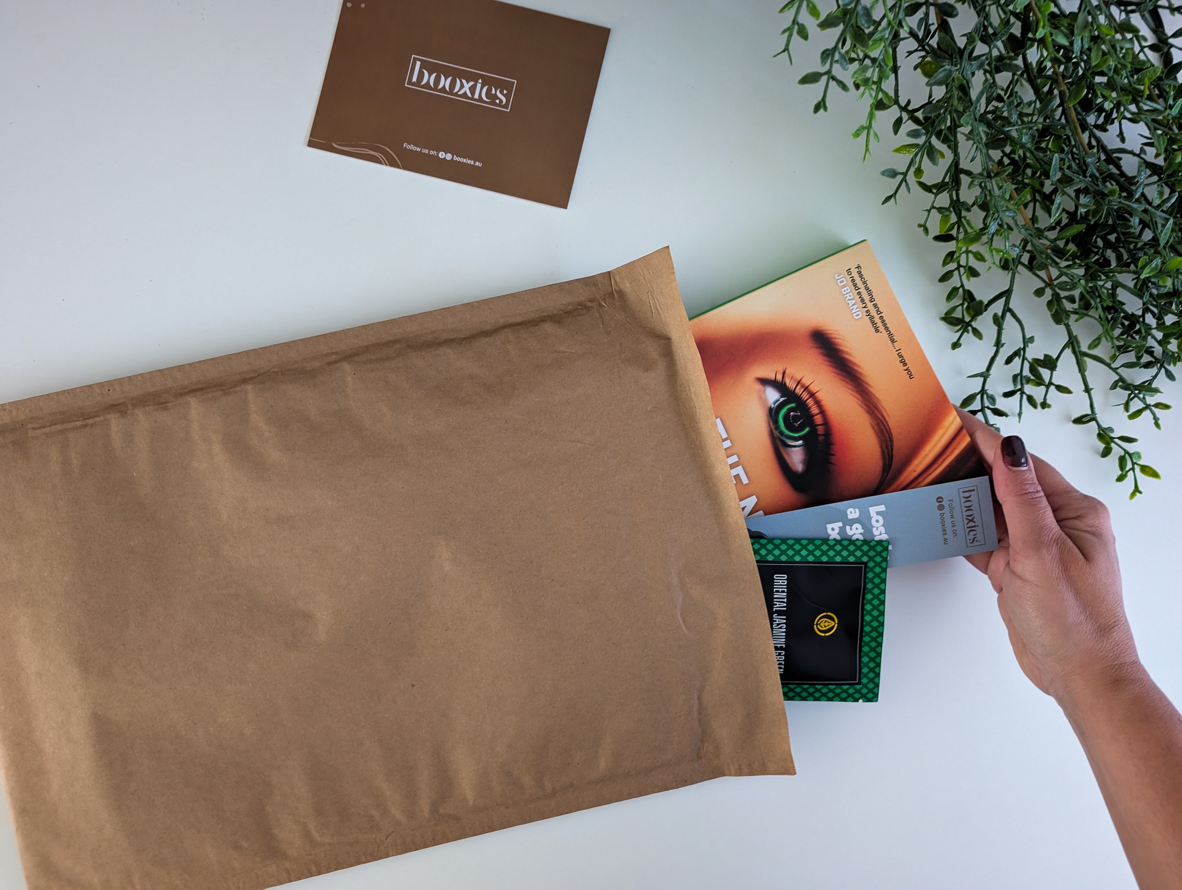 Person holding a product package next to a brown envelope on a white surface with a plant in the background.