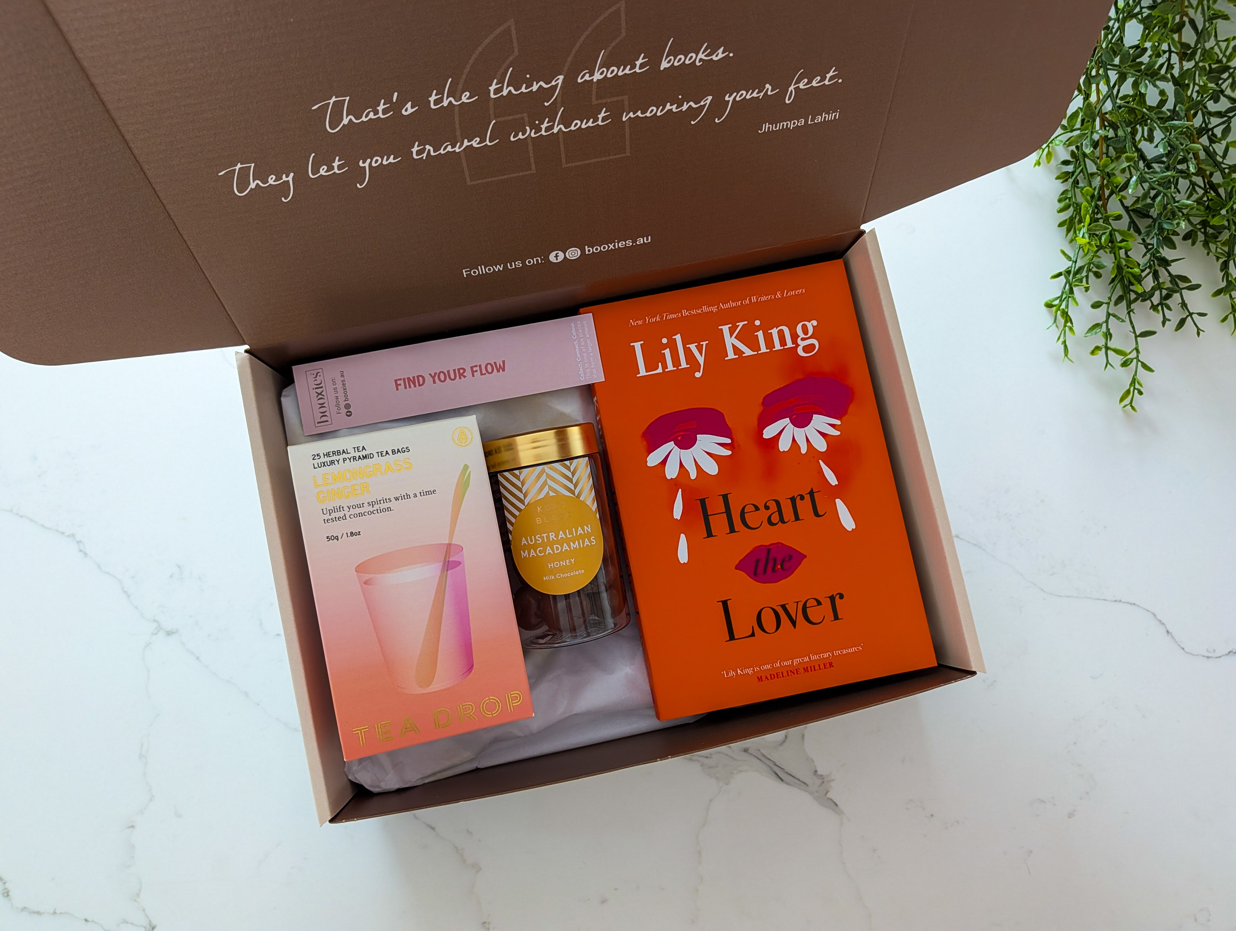 Open box containing a book titled 'Heart Lover' by Lily King, a small jar, and other items on a white surface.