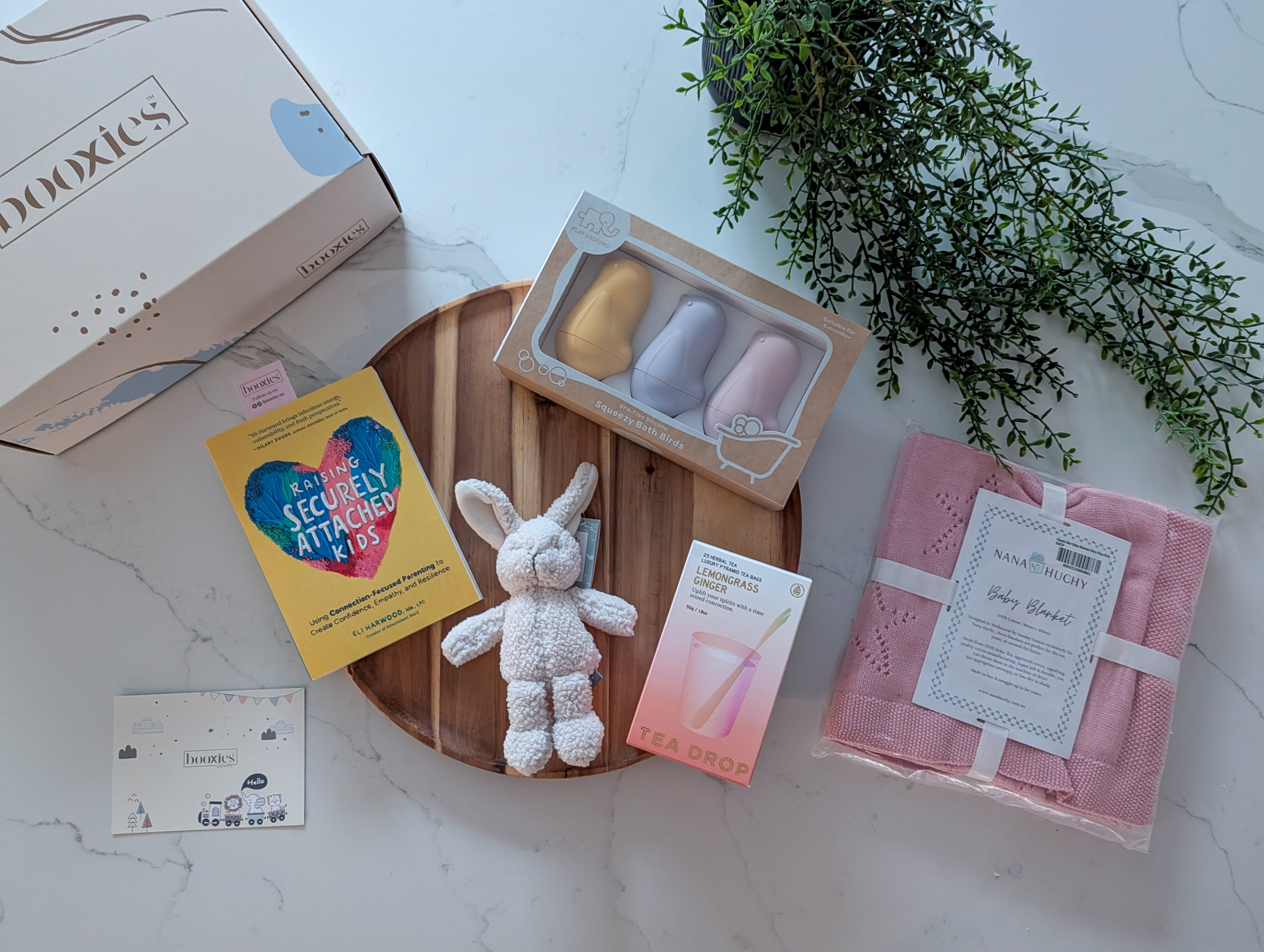 Collection of baby items including a toy, cards, and a blanket on a white surface with a plant in the background.