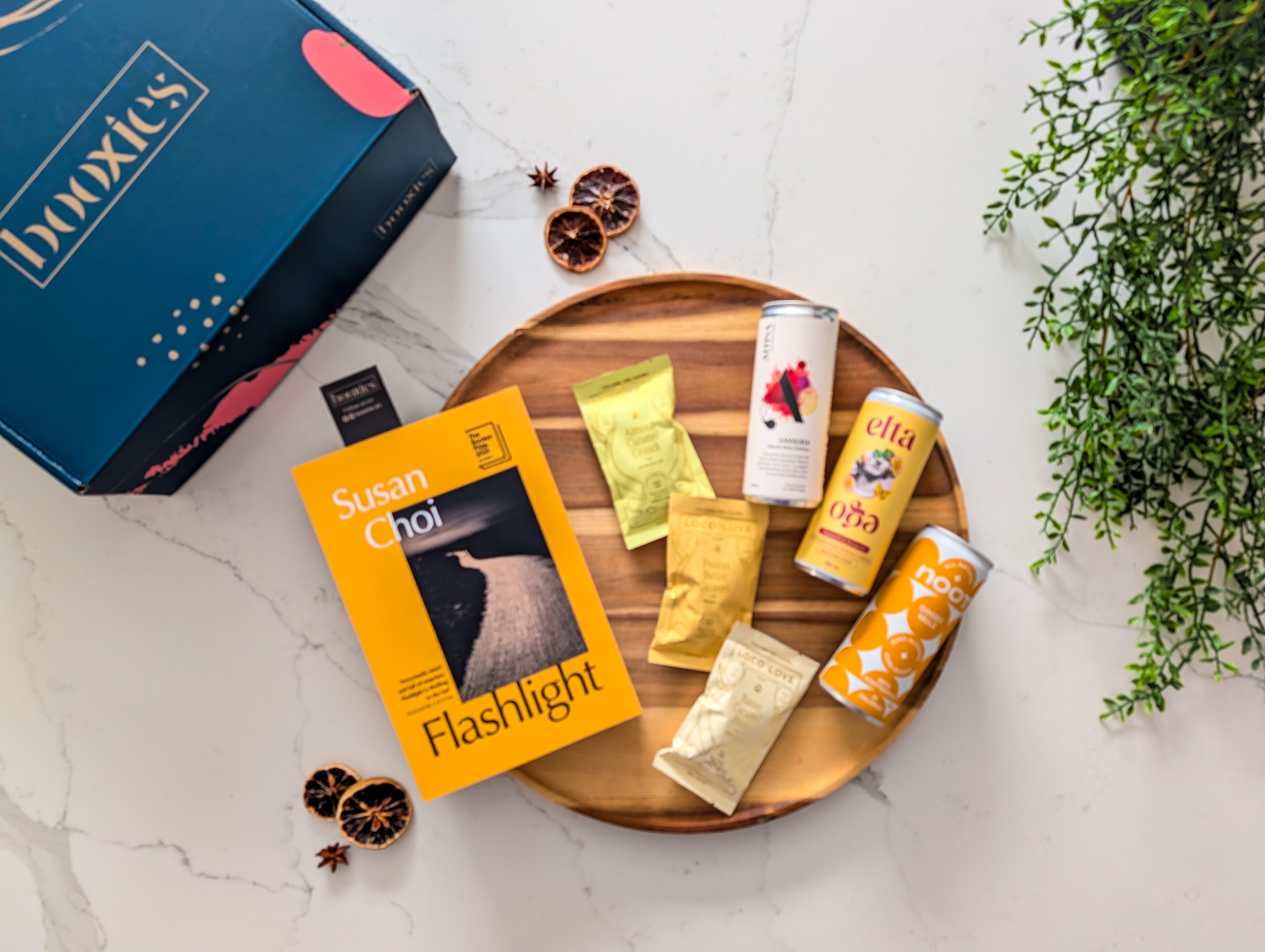 Assorted loco love chocolate and non-alcoholic drinks on a wooden tray with a 'booxies' branded box and a new book in the background. making up the Zero Edit curated booxies