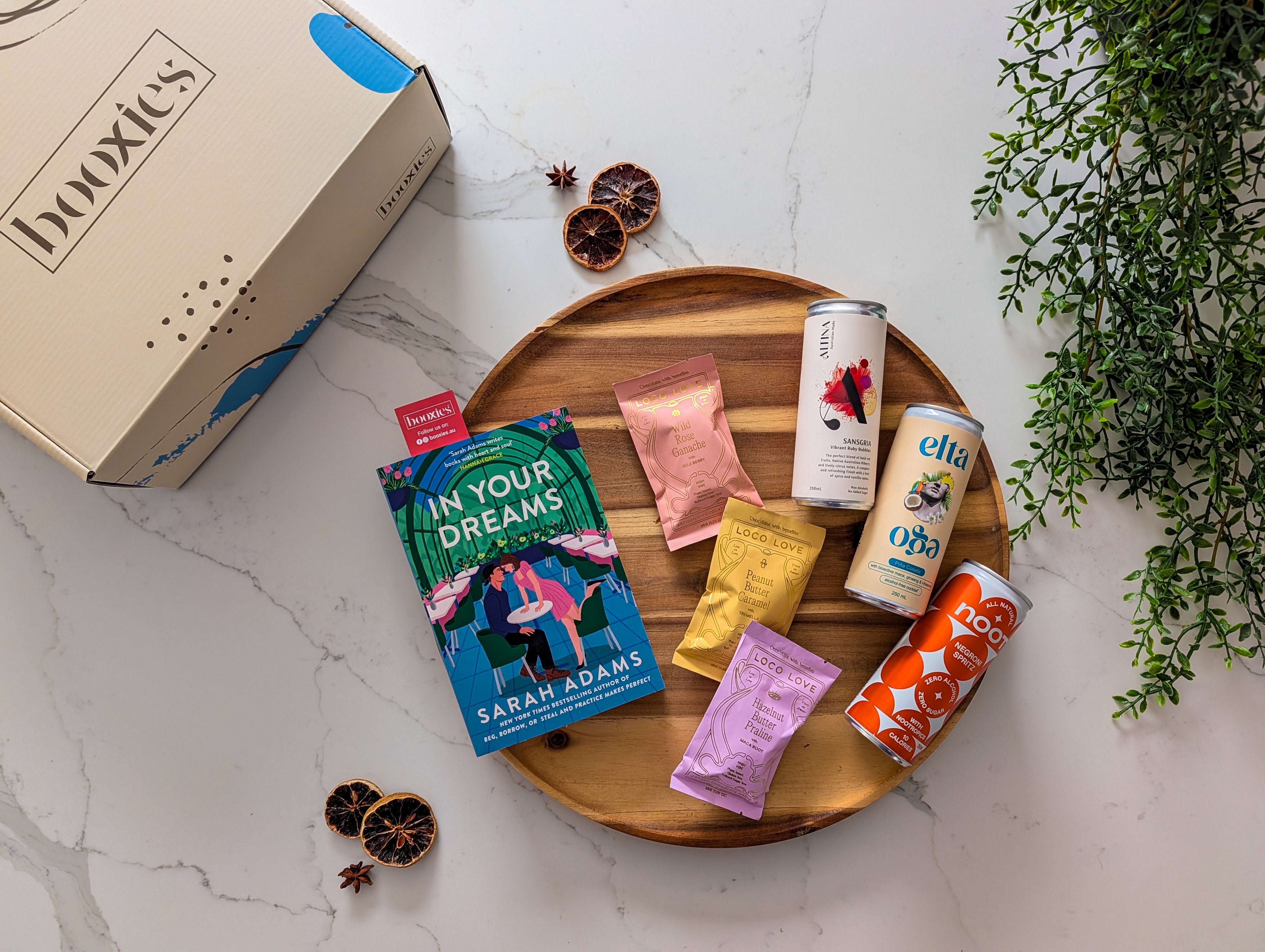 Assorted loco love chocolate and non-alcoholic drinks on a wooden tray with a 'booxies' branded box and a new book in the background. making up the Zero Edit curated booxies