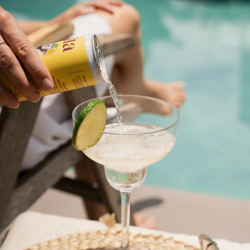 Person pouring a drink from a can into a glass with a lime slice, by a pool.